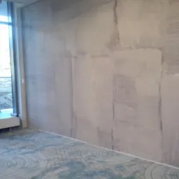 Ściana z efektem dekoracyjnym imitującym kamień, widoczne niedoskonałości i różnice w odcieniach szarości, na podłodze wzorzysta wykładzina w odcieniach błękitu i szarości, widoczne oświetlenie...