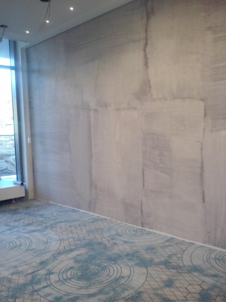 Ściana z efektem dekoracyjnym imitującym kamień, widoczne niedoskonałości i różnice w odcieniach szarości, na podłodze wzorzysta wykładzina w odcieniach błękitu i szarości, widoczne oświetlenie...