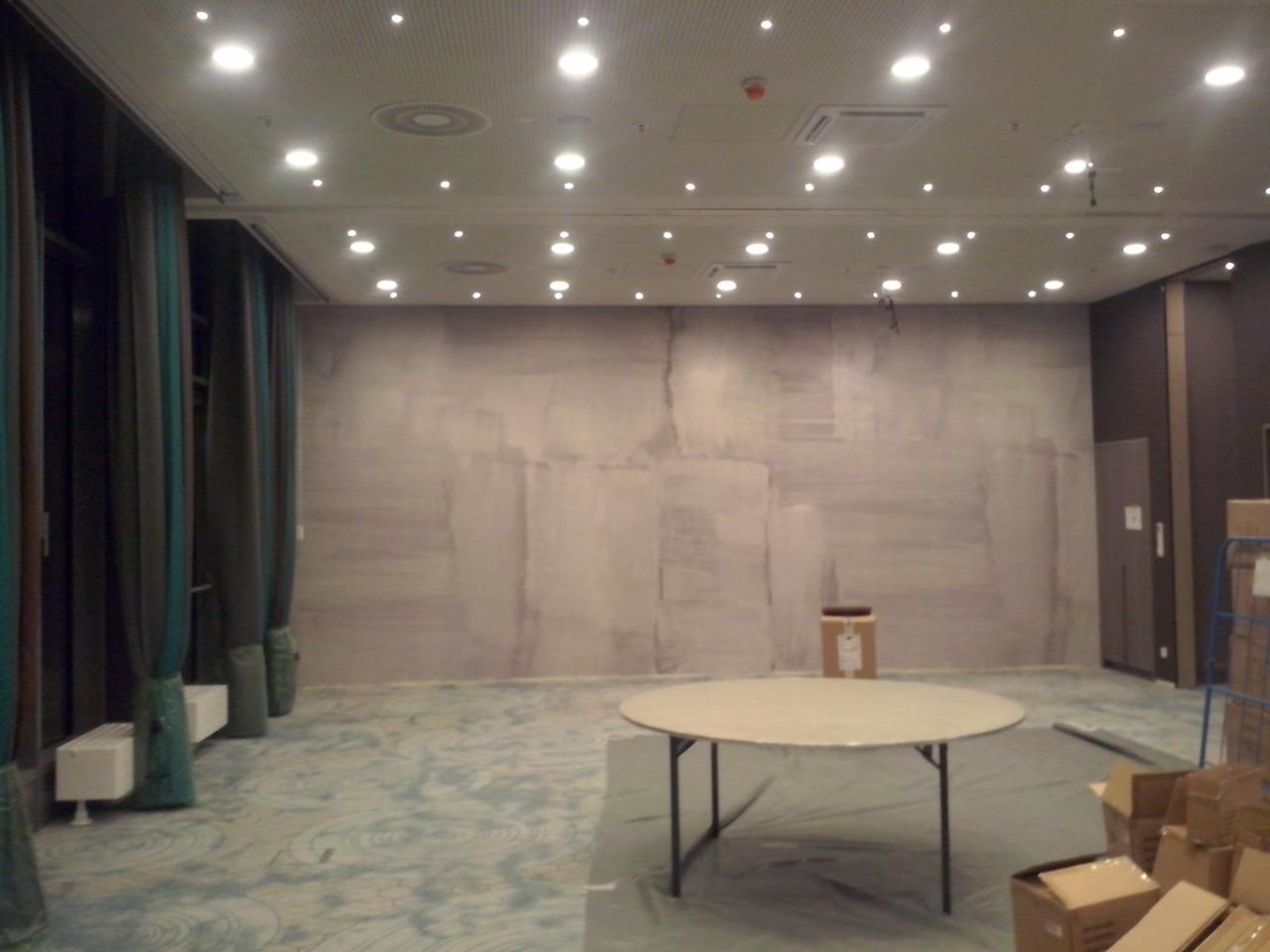 Duże pomieszczenie biurowe w trakcie szpachlowania ścian, widoczne ślady po szpachli, okrągły stół na środku, kartony w rogu, zasłonięte grzejniki, oświetlenie sufitowe.