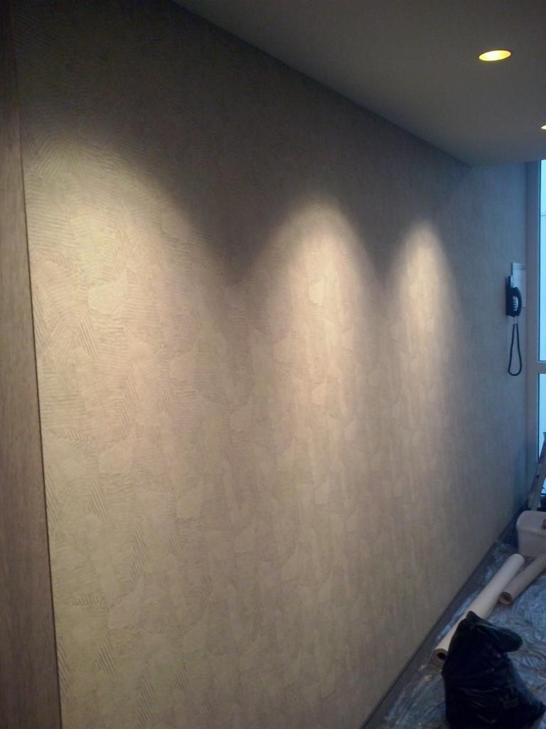 Ściana z nową, teksturowaną tapetą w odcieniach beżu, oświetlona punktowo, widoczne rolki tapety i worek na ścinki przy podłodze.