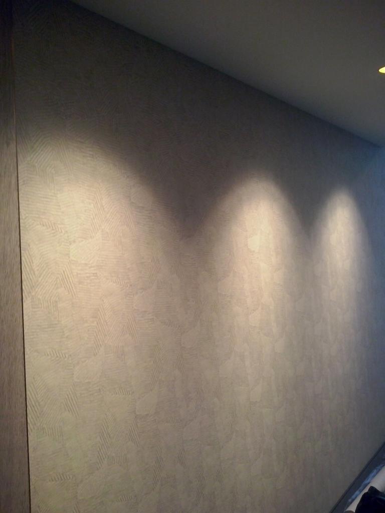 Ściana pokryta tapetą z delikatnym geometrycznym wzorem w odcieniach szarości, oświetlona trzema punktowymi lampami sufitowymi, ukazującymi fakturę materiału.