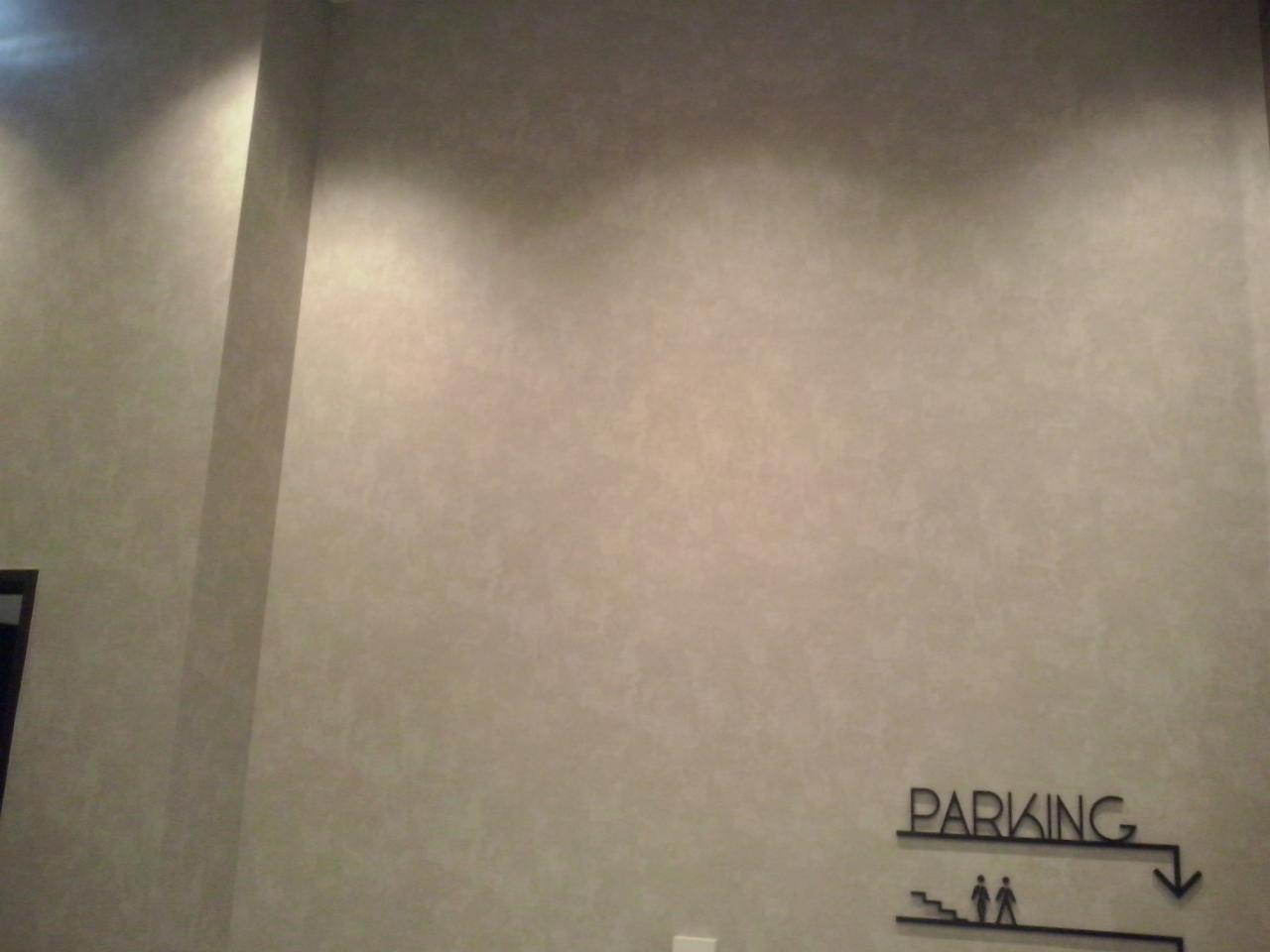 Ściana w biurze pomalowana na jasny, beżowy kolor z delikatną fakturą, widoczny znak kierunkowy 'PARKING' z piktogramem schodów i sylwetkami ludzi, oświetlenie punktowe tworzy gradient na górze...