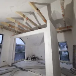 Poddasze w trakcie adaptacji: widoczne drewniane belki stropowe zabezpieczone folią, ściany przygotowane pod malowanie, okna dachowe w czarnych ramach, przewody elektryczne na podłodze.