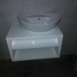 Biała szafka łazienkowa z półkami pod umywalką nablatową w trakcie montażu, widoczna poziomica i niedokończone wykończenie podłogi.