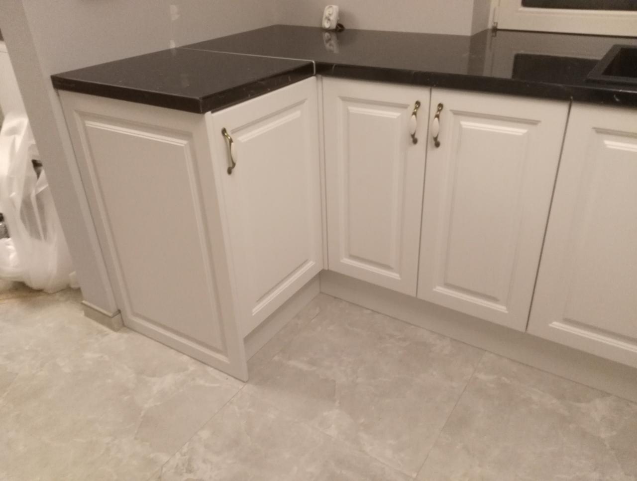 Fragment zabudowy kuchennej: białe szafki z frezowanymi frontami i ciemny, nakrapiany blat. Widoczne porcelanowe uchwyty z metalowym wykończeniem.