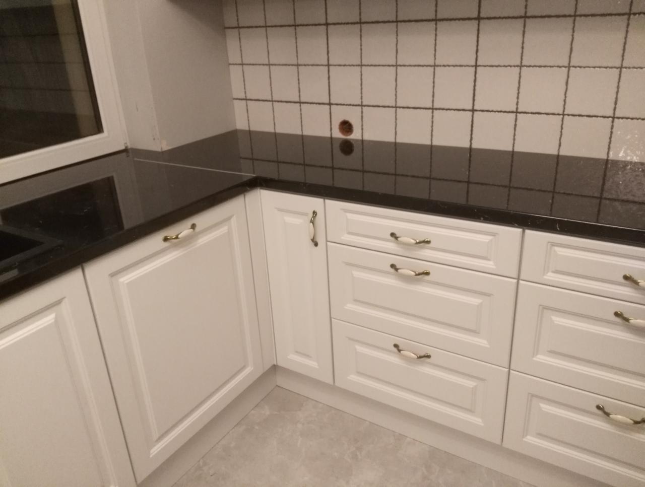 Białe szafki kuchenne z frezowanymi frontami i czarnym blatem, kremowe uchwyty, ściana wyłożona kwadratowymi płytkami.