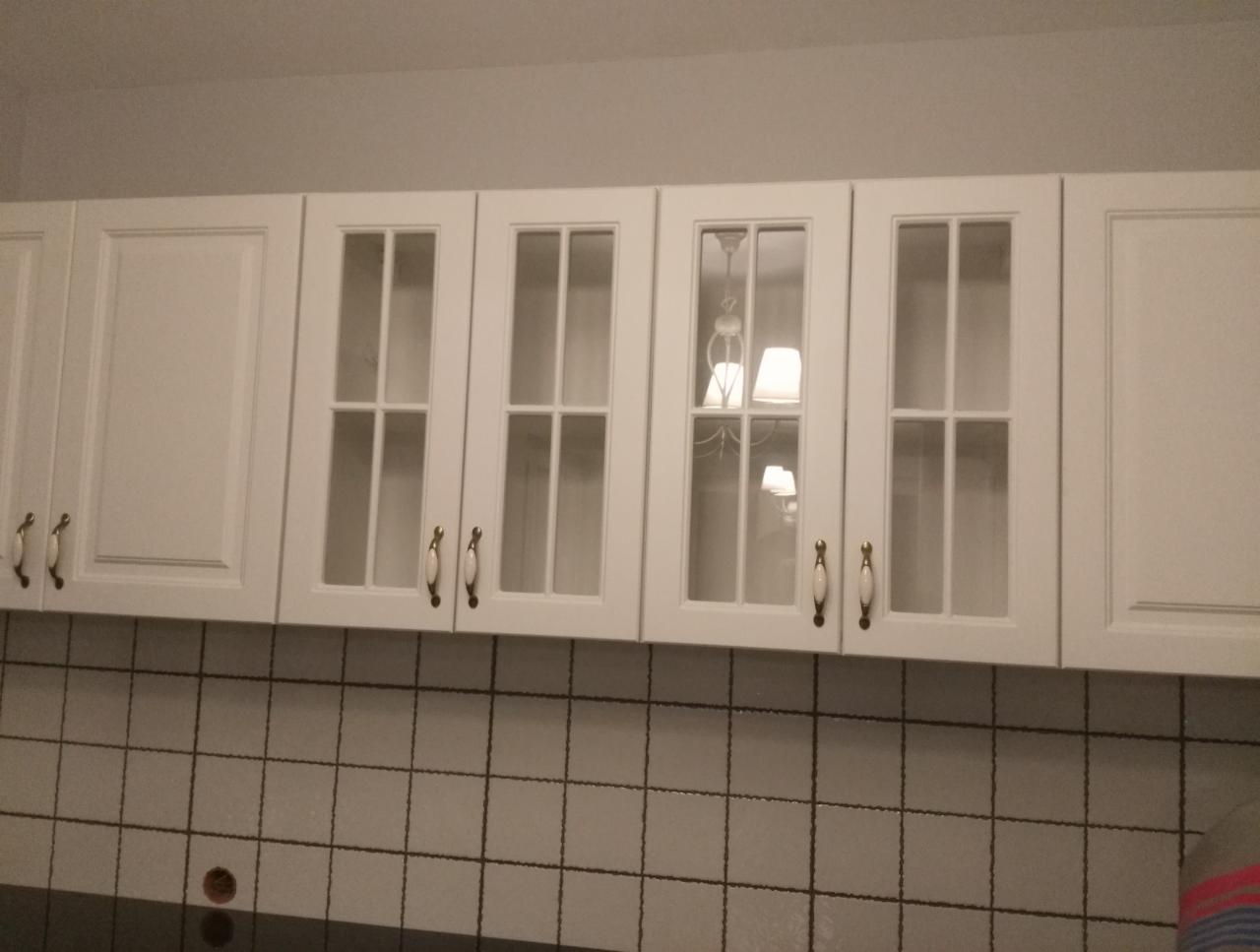 Białe szafki kuchenne wiszące z przeszklonymi frontami i uchwytami w stylu retro, widoczne odbicie lampy w szkle, tło z płytek w kratkę.