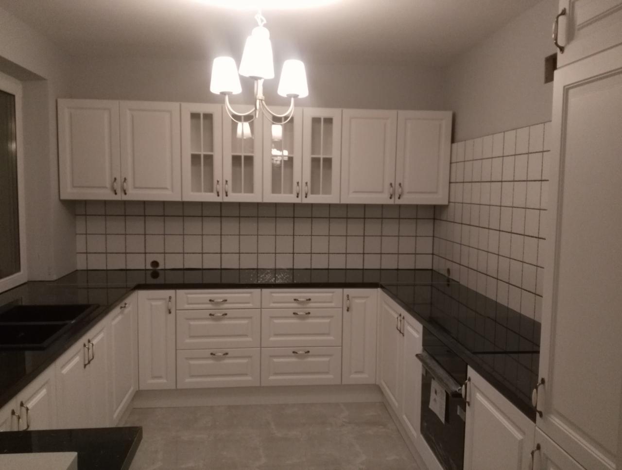 Nowoczesna kuchnia z białymi szafkami i czarnym blatem, oświetlona wiszącą lampą z białymi kloszami, z widocznymi białymi płytkami na ścianie.