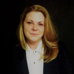 Portret kobiety w białej koszuli i ciemnej marynarce, z blond włosami, uśmiechająca się do kamery na ciemnym tle.