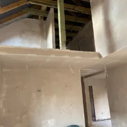 Surowe wnętrze w trakcie prac wykończeniowych: świeżo nałożone tynki maszynowe na ścianach i suficie, widoczne wystające kable elektryczne, drewniana konstrukcja dachu, plastikowe krzesła i butelka...