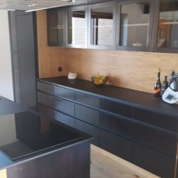 Stolarstwo Zakład PHU Jarosław Jurek - Nowoczesna kuchnia z czarnymi frontami szafek i drewnianym panelem ściennym. Na blacie oświetlonym od góry stoją owoce i akcesoria kuchenne.