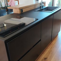 Stolarstwo Zakład PHU Jarosław Jurek - Czarna wyspa kuchenna z płytą indukcyjną i zlewem, drewniany blat barowy, jasna podłoga z desek, nowoczesny design, widok z boku, naturalne światło.