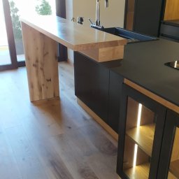 Stolarstwo Zakład PHU Jarosław Jurek - Nowoczesna kuchnia z drewnianym blatem i wyspą. Minimalistyczny design, czarne szafki z oświetleniem LED, drewniana podłoga. Widok na okno z zielenią w tle.
