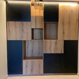 Stolarstwo Zakład PHU Jarosław Jurek - Nowoczesna szafa wnękowa z elementami drewnopodobnymi i czarnymi, oświetlona od góry, z otwartymi półkami i asymetrycznym układem paneli.