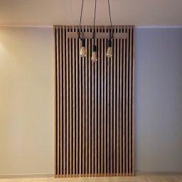 Stolarstwo Zakład PHU Jarosław Jurek - Drewniana ażurowa ścianka działowa z listew, podwieszona pod sufitem, oświetlona trzema żarówkami na czarnych kablach. Minimalistyczny design, jasne ściany w tle.