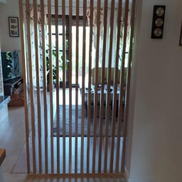 Stolarstwo Zakład PHU Jarosław Jurek - Drewniana ścianka działowa z pionowych listew, oddzielająca przestrzeń w jasnym wnętrzu. Widoczne elementy wyposażenia salonu w tle.