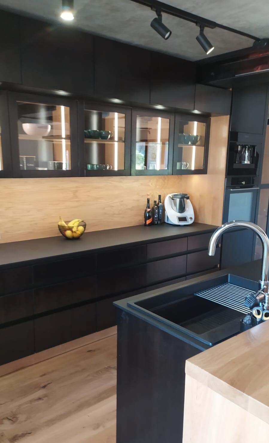 Nowoczesna, czarna zabudowa kuchenna z podświetlanymi szafkami i drewnianym blatem. Na blacie owoce, butelki i robot kuchenny. Minimalistyczny design, oświetlenie punktowe.