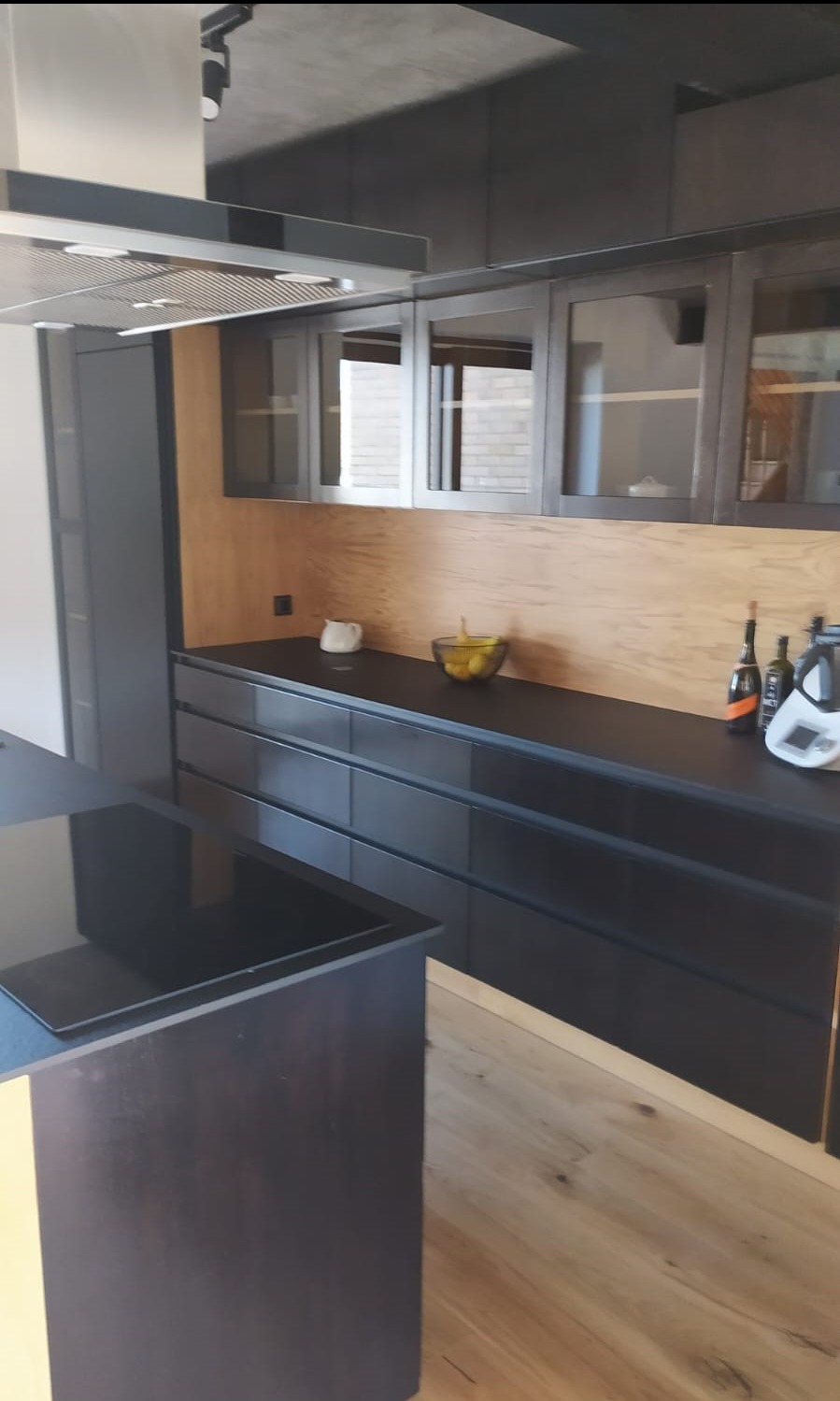 Nowoczesna kuchnia z czarnymi frontami szafek i drewnianym panelem ściennym. Na blacie oświetlonym od góry stoją owoce i akcesoria kuchenne.