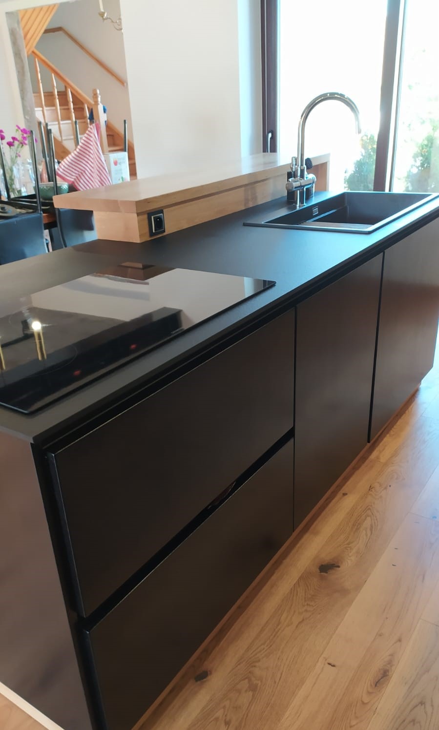 Czarna wyspa kuchenna z płytą indukcyjną i zlewem, drewniany blat barowy, jasna podłoga z desek, nowoczesny design, widok z boku, naturalne światło.