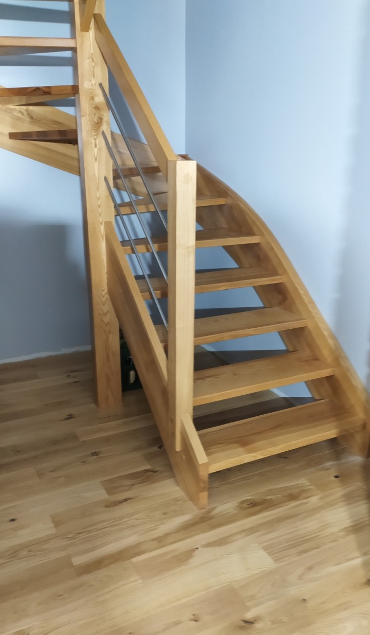 Jasne, drewniane schody z metalową balustradą, zakręcające w lewo, na tle błękitnej ściany. Widok z dołu, ukazujący strukturę drewna i minimalistyczny design.