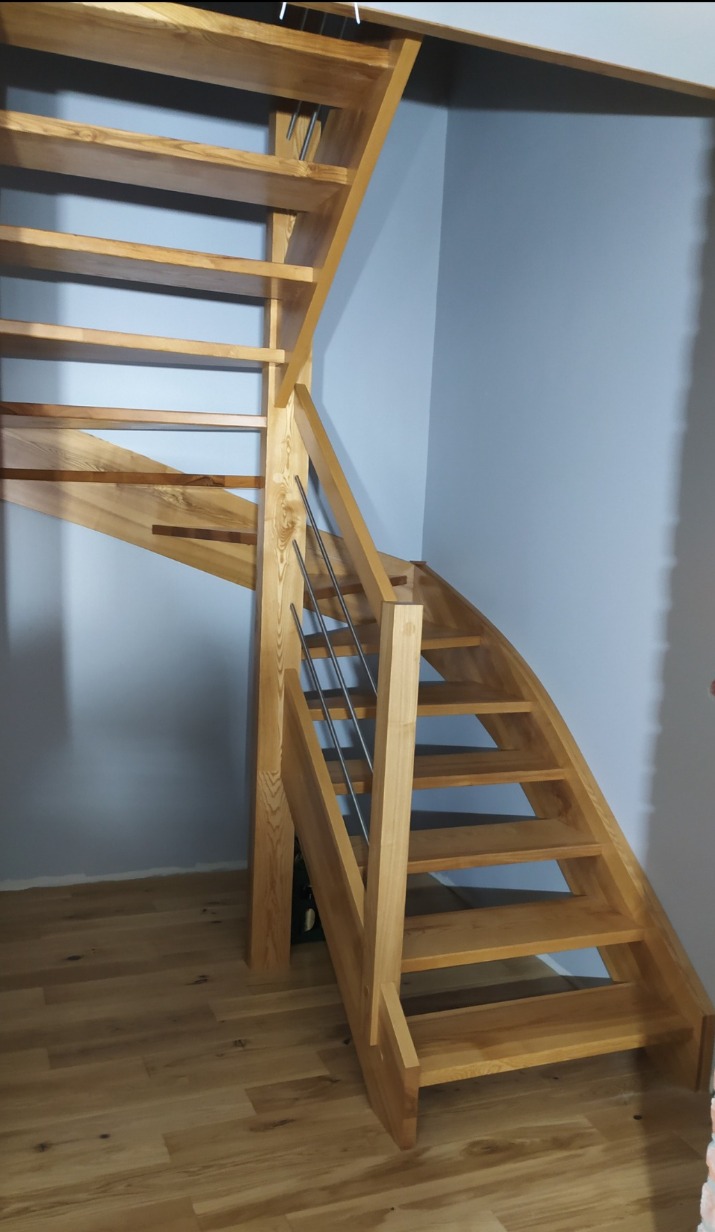 Drewniane schody kręcone z metalową balustradą, wykonane na zamówienie, z widocznym usłojeniem drewna i naturalnym odcieniem, na tle szarej ściany i drewnianej podłogi.