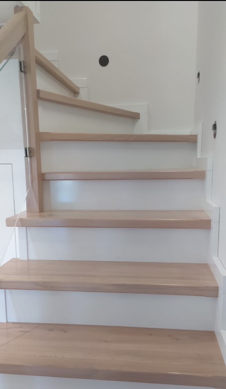 Jasne, drewniane stopnie schodów z białymi podstopnicami i szklaną balustradą. Minimalistyczny design, widok z dołu, oświetlenie naturalne.