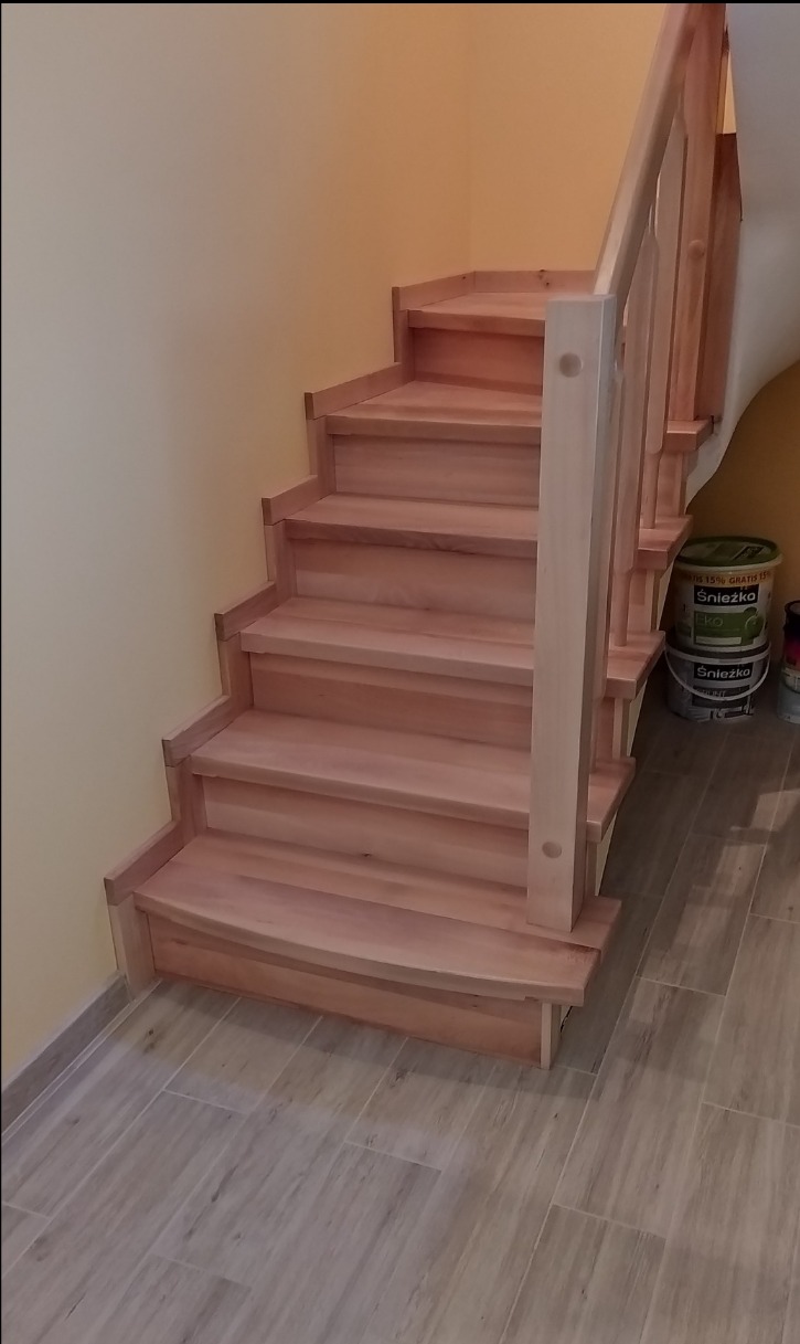 Jasne, drewniane schody z balustradą, prowadzące w górę. Widoczne detale wykończenia i naturalna kolorystyka drewna. W tle ściana w odcieniach beżu i pojemniki z farbą.