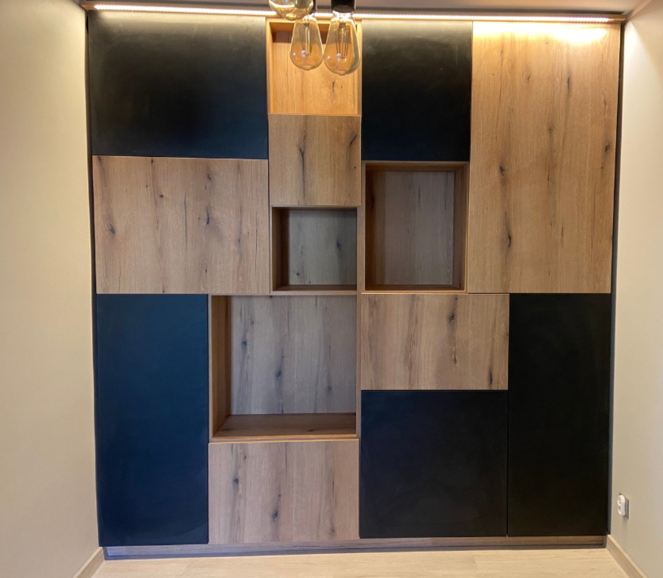 Nowoczesna szafa wnękowa z elementami drewnopodobnymi i czarnymi, oświetlona od góry, z otwartymi półkami i asymetrycznym układem paneli.