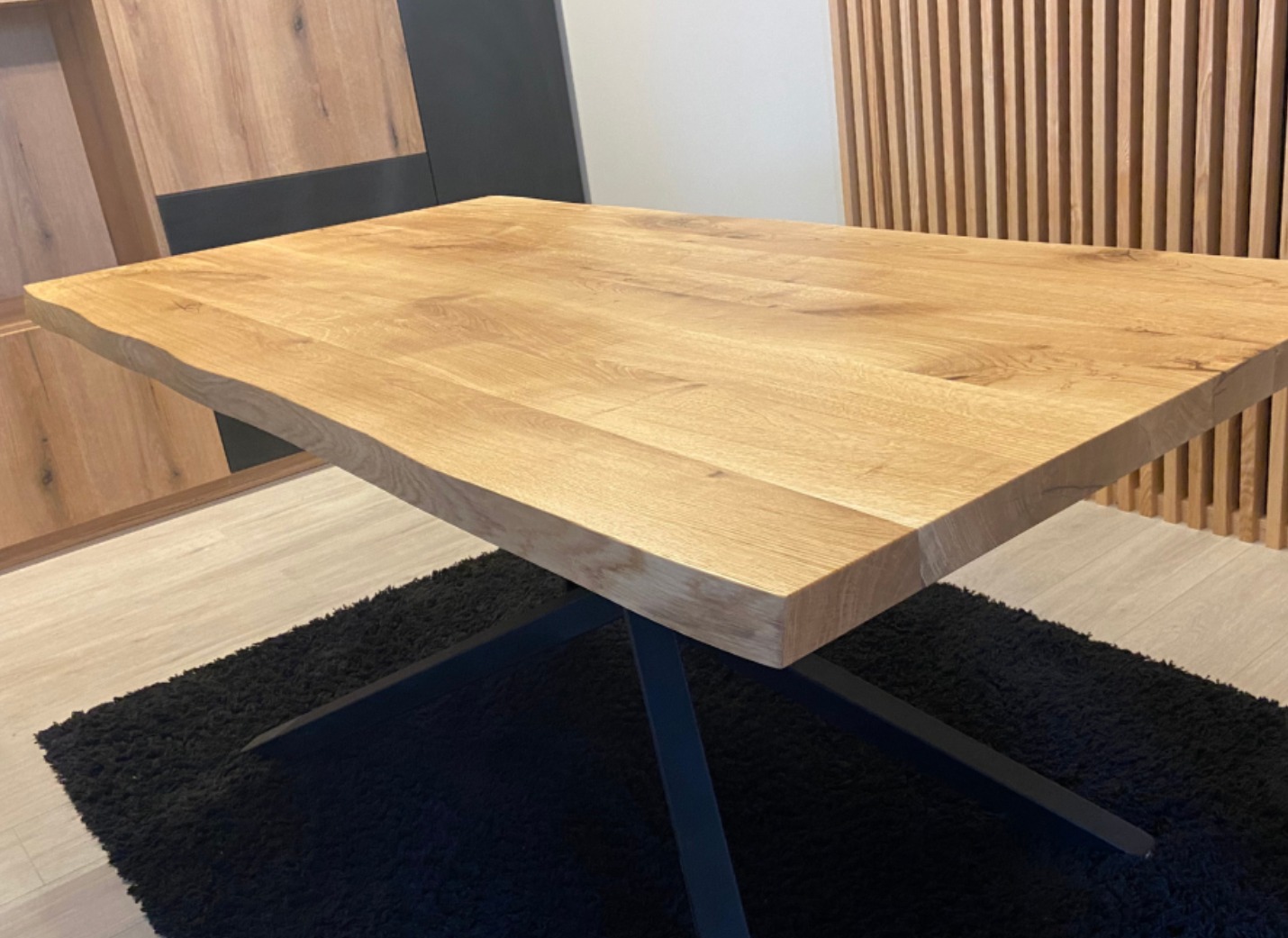 Stół z blatem z jasnego drewna o nieregularnej krawędzi, wsparty na czarnej, metalowej podstawie. W tle drewniana ściana z pionowych listew i fragment szafki.