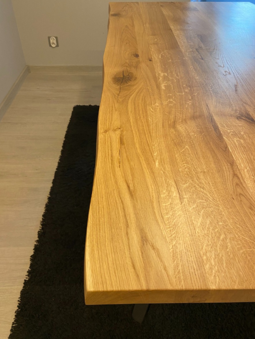 Jasny, dębowy blat stołu z wyraźnym usłojeniem i naturalną krawędzią, ustawiony na tle czarnego dywanu i jasnej podłogi. Widoczny fragment gniazdka elektrycznego.