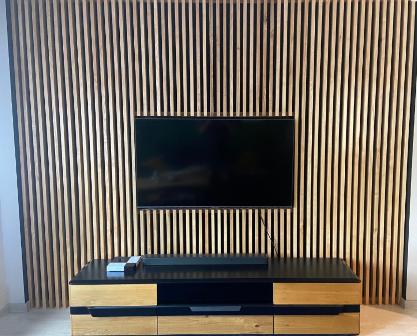 Drewniana ściana z pionowych listew za telewizorem i szafką RTV. Naturalne drewno, nowoczesny design, minimalistyczny styl. Widoczne kable.