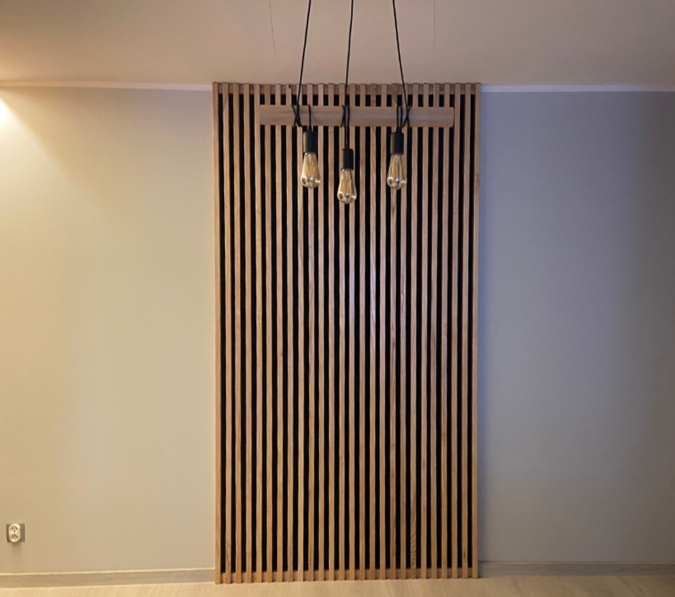 Drewniana ażurowa ścianka działowa z listew, podwieszona pod sufitem, oświetlona trzema żarówkami na czarnych kablach. Minimalistyczny design, jasne ściany w tle.
