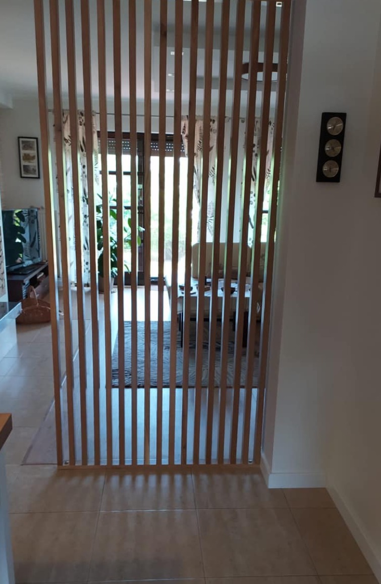 Drewniana ścianka działowa z pionowych listew, oddzielająca przestrzeń w jasnym wnętrzu. Widoczne elementy wyposażenia salonu w tle.