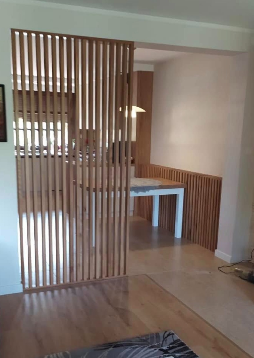 Drewniana ścianka działowa z pionowych listew oddziela przestrzeń jadalni. Widoczny stół z białymi nogami i drewnianym blatem oraz fragment drewnianej boazerii.