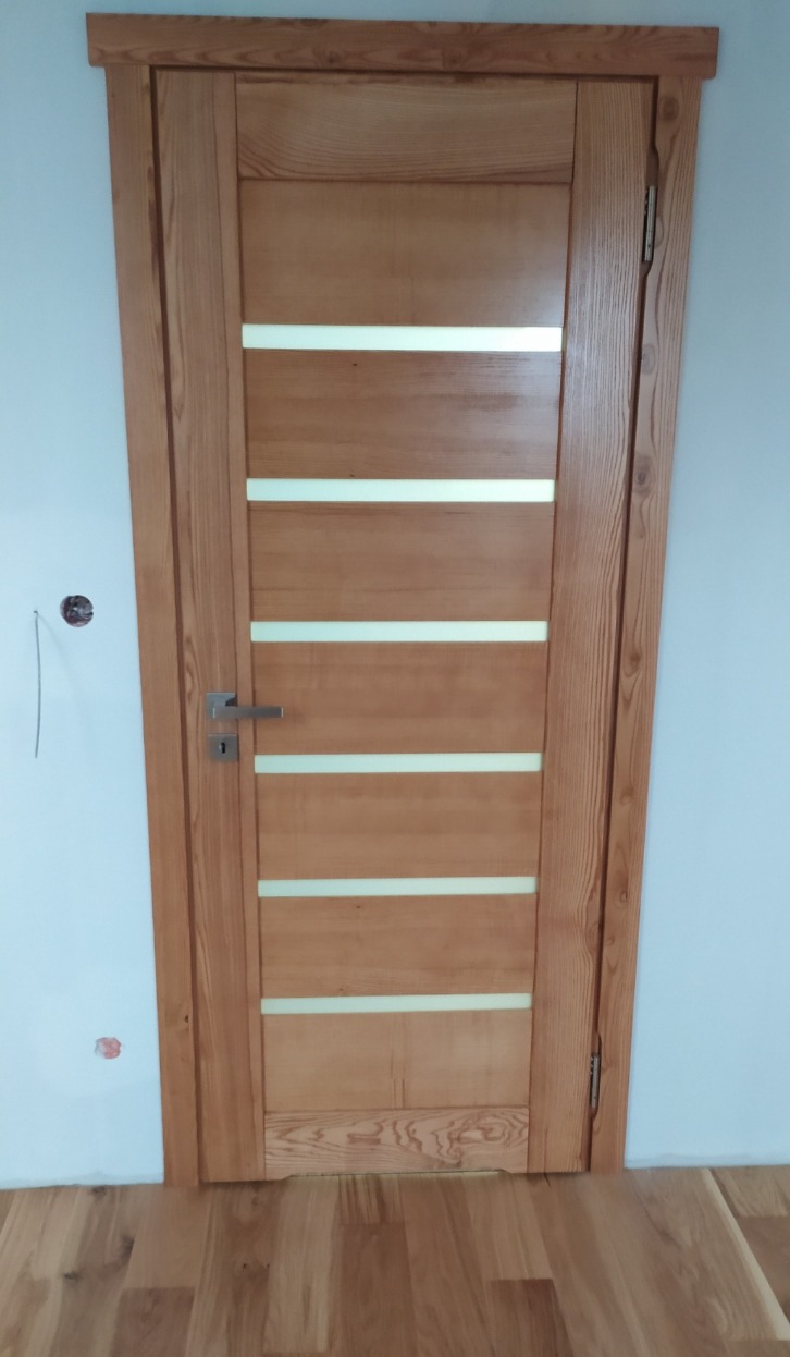 Jasne, drewniane drzwi wewnętrzne z poziomymi przeszkleniami, widoczne na tle białej ściany i drewnianej podłogi. Klamka po lewej stronie.