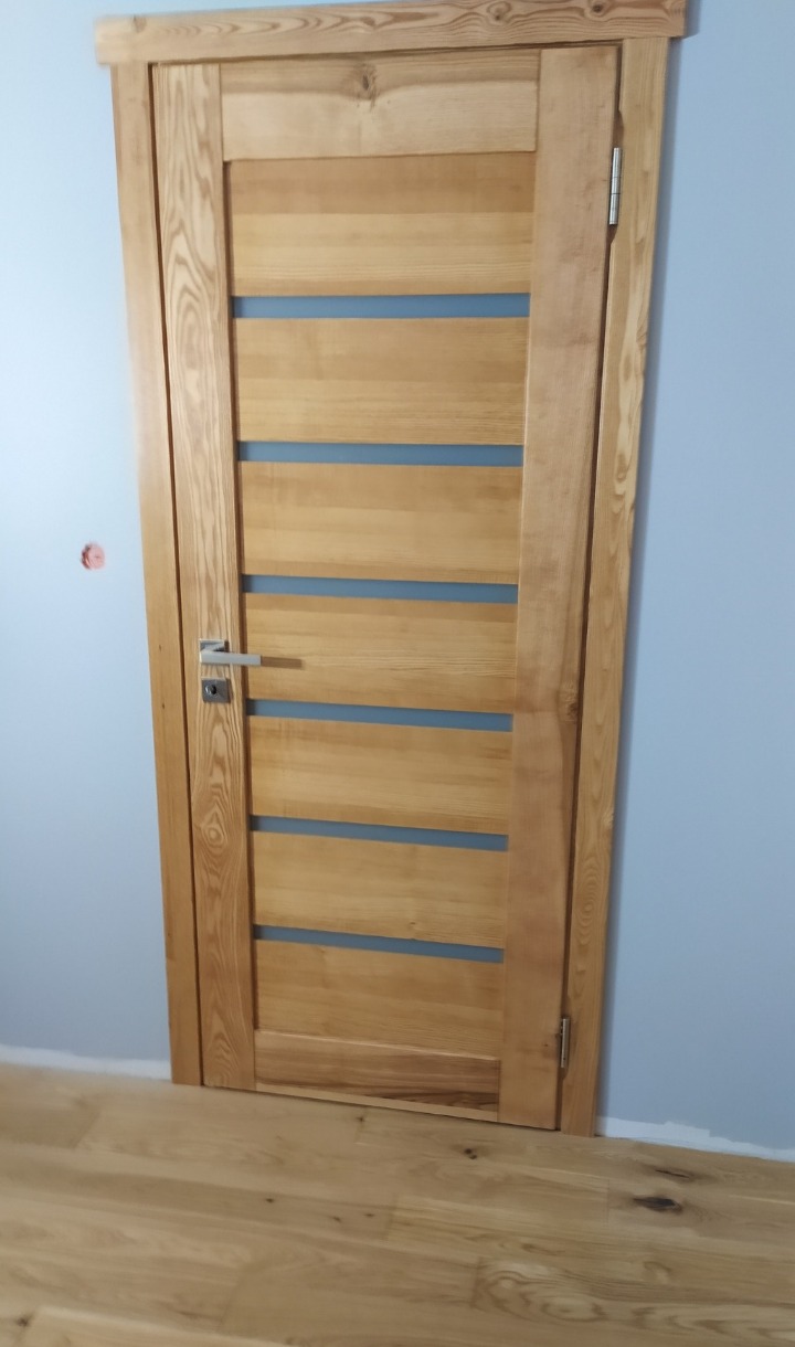 Jasne, drewniane drzwi wewnętrzne z poziomymi przeszkleniami, widoczne na tle błękitnej ściany i drewnianej podłogi. Minimalistyczny design, naturalne wykończenie.