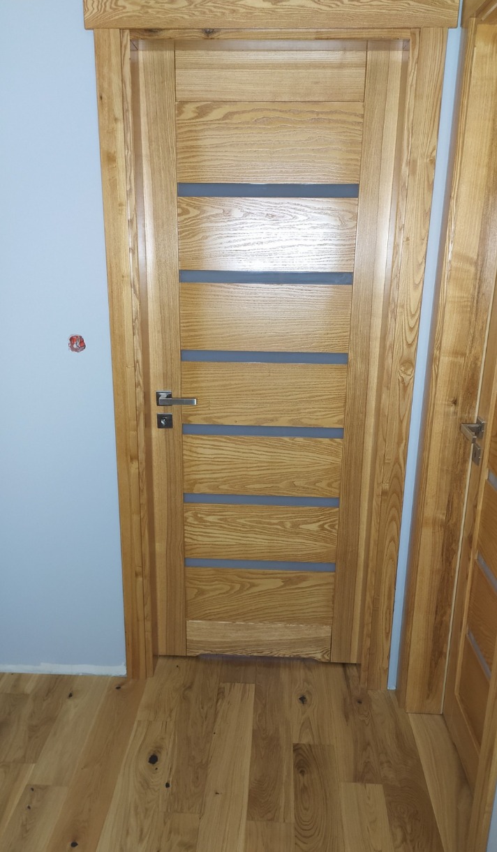 Nowoczesne drzwi z drewna jesionowego z przeszkleniami, zamontowane w Chodzieży. Naturalne wykończenie, widoczna struktura drewna, srebrna klamka.