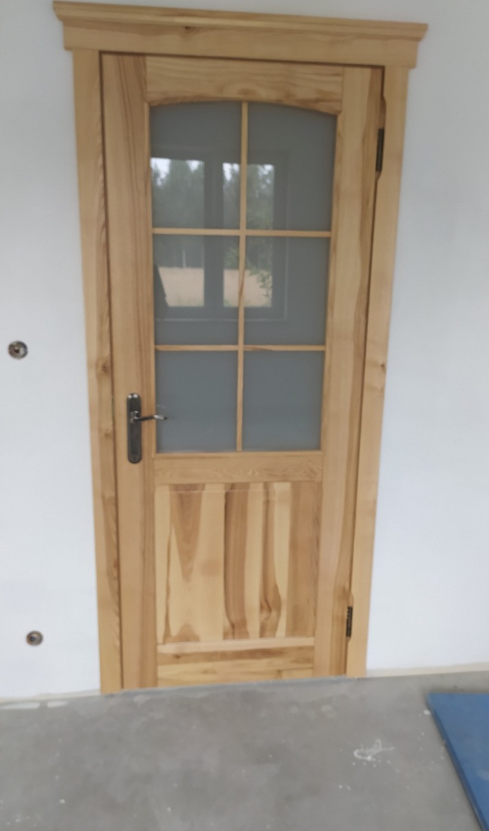 Jasne, przeszklone drzwi z naturalnego drewna, z widoczną strukturą słojów, zamontowane w białej ścianie. Widoczne detale wykończenia i klamka.