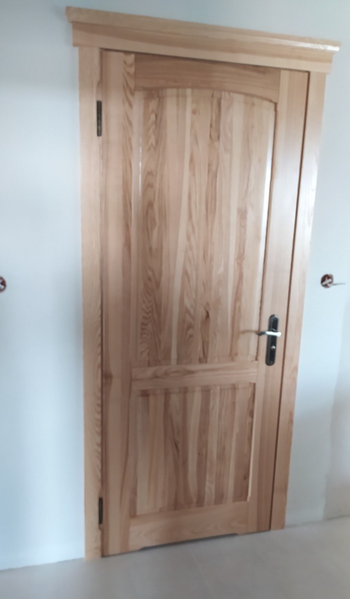Jasne, drewniane drzwi wewnętrzne z ozdobną ramą i panelem. Widoczne zawiasy i klamka. Świeżo zamontowane, w surowym wnętrzu.