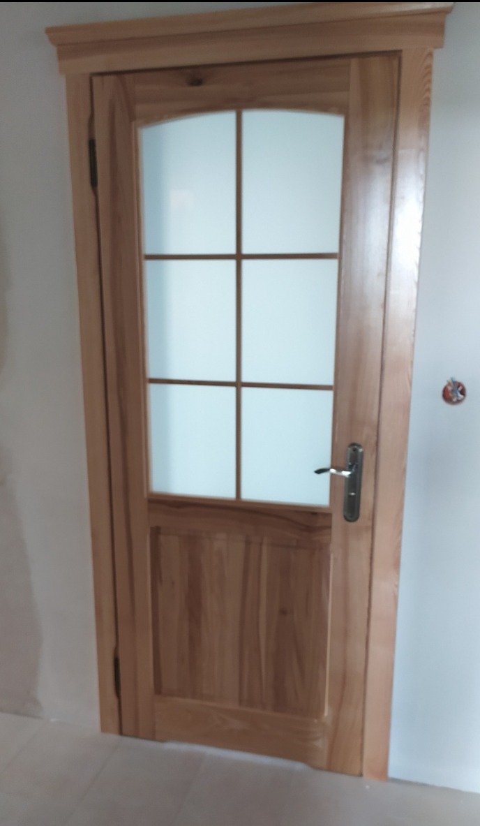 Jasne, drewniane drzwi wewnętrzne z szybą w kratkę, widoczne zawiasy i klamka. Minimalistyczny design, ciepłe oświetlenie, surowy stan wykończenia wnętrza.