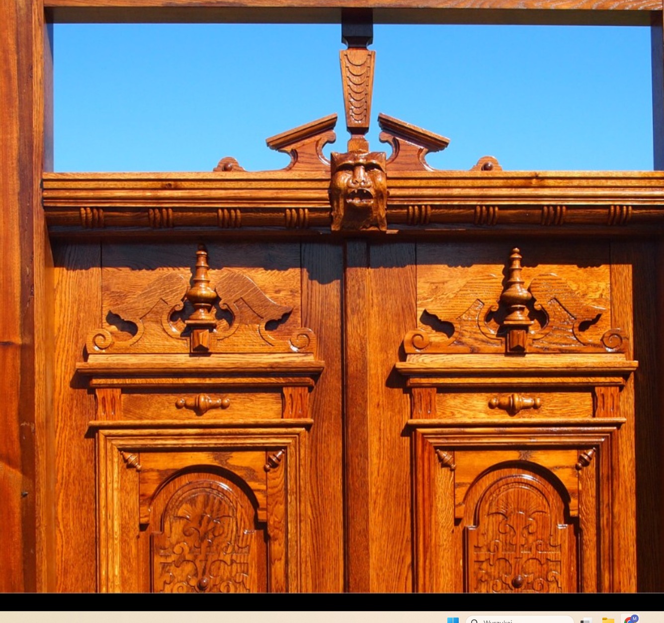Bogato zdobione, dwuskrzydłowe drzwi z rzeźbionego drewna w ciepłym odcieniu brązu, zwieńczone motywem maski na tle błękitnego nieba.