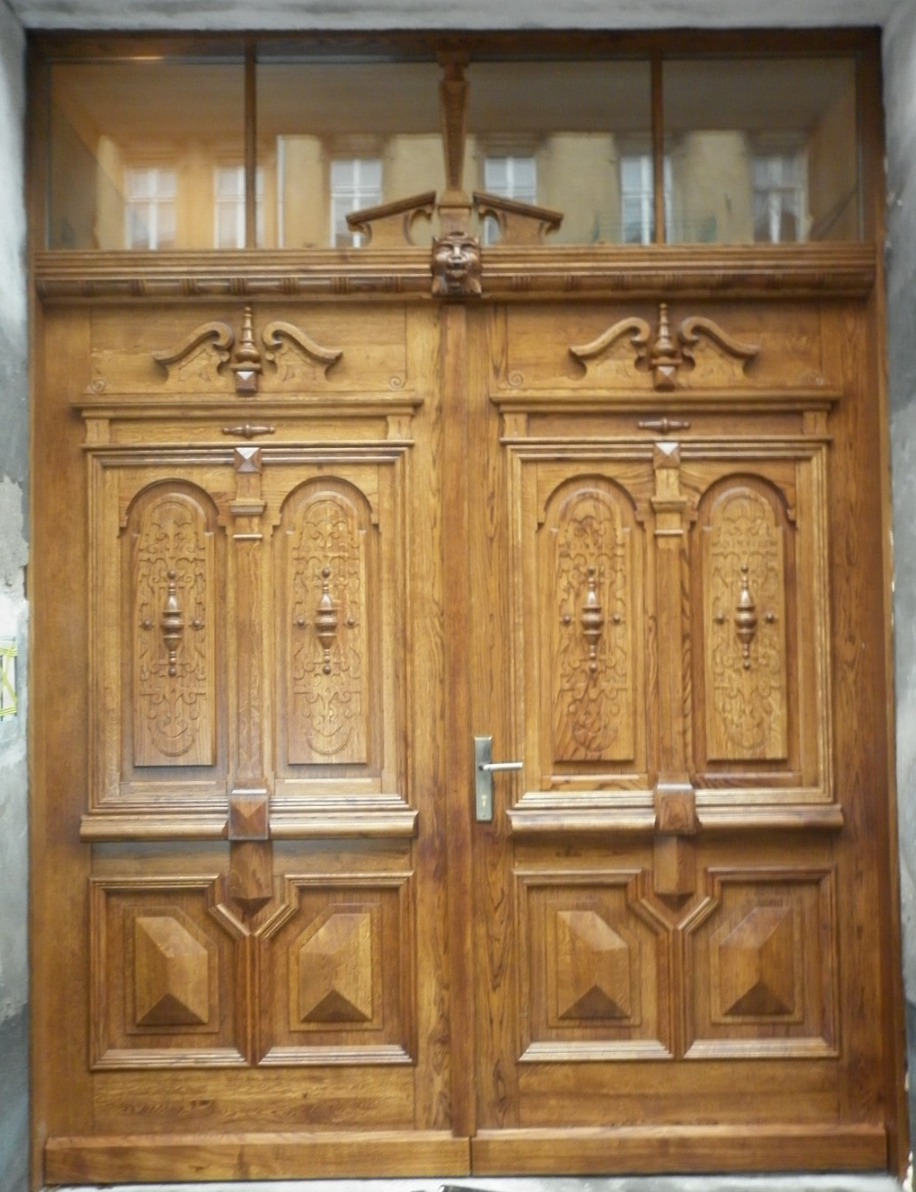 Dwuskrzydłowe, zdobione drzwi z litego drewna o ciepłym odcieniu, zwieńczone rzeźbionym ornamentem i przeszkleniem, ukazujące wnętrze budynku.