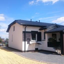 Zakład Ogólnobudowlany ZEN- BUD Zenon Kuźnik - Wykończony dom jednorodzinny z białą elewacją, czarnym dachem i elementami dekoracyjnymi z drewna, otoczony jasnym żwirem i brukowanym podjazdem w słoneczny dzień.