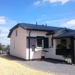 Wykończony dom jednorodzinny z białą elewacją, czarnym dachem i elementami dekoracyjnymi z drewna, otoczony jasnym żwirem i brukowanym podjazdem w słoneczny dzień.