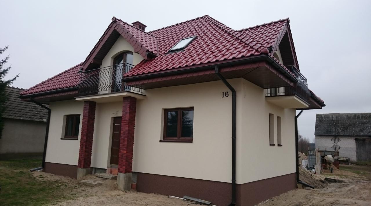 Dwupiętrowy dom jednorodzinny z czerwonym dachem, beżową elewacją i balkonami z czarną balustradą, widok z zewnątrz w pochmurny dzień.