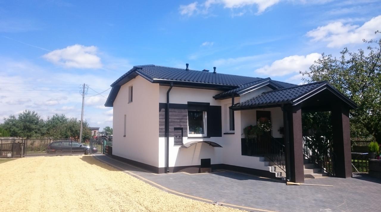 Wykończony dom jednorodzinny z białą elewacją, czarnym dachem i elementami dekoracyjnymi z drewna, otoczony jasnym żwirem i brukowanym podjazdem w słoneczny dzień.