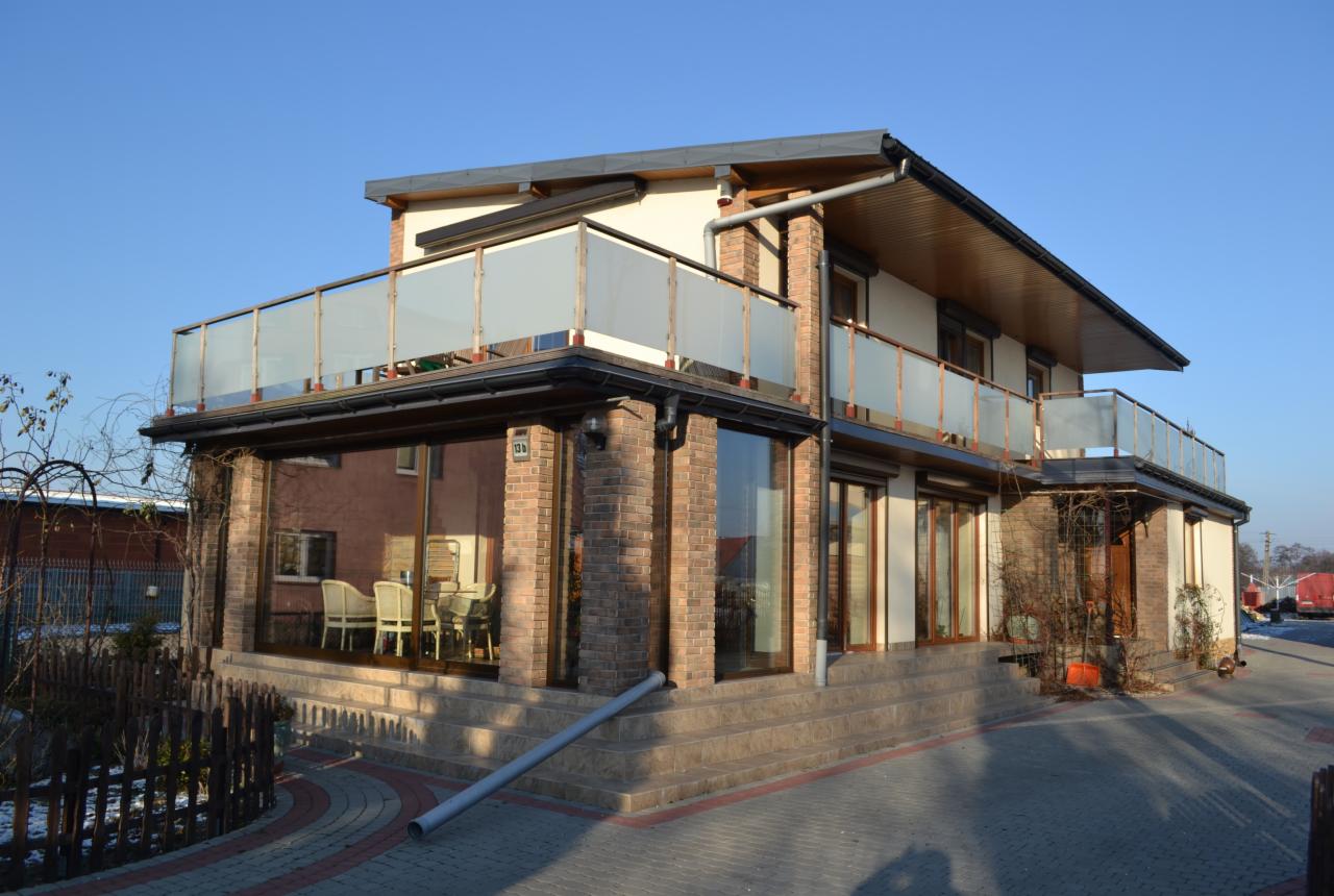 Dwupiętrowy dom z jasną elewacją, balkonami z mlecznego szkła i ceglanymi elementami wykończeniowymi, widoczne schody wejściowe i fragment posesji