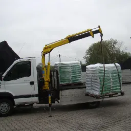 Biała ciężarówka z żółtym dźwigiem podnosi paletę z workami na tle hałdy węgla