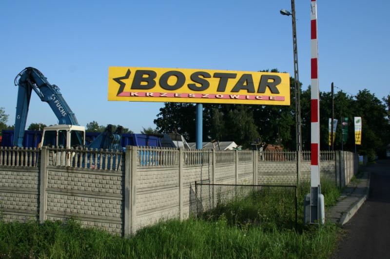 Tablica reklamowa z nazwą 'BOSTAR KRZESZOWICE' na tle błękitnego nieba, widoczny ciężki sprzęt budowlany za betonowym ogrodzeniem i słup z biało-czerwonymi pasami.