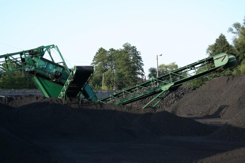 Zielony podajnik taśmowy nad stertą czarnego węgla na składowisku, widok z dołu, w tle drzewa i słup oświetleniowy.